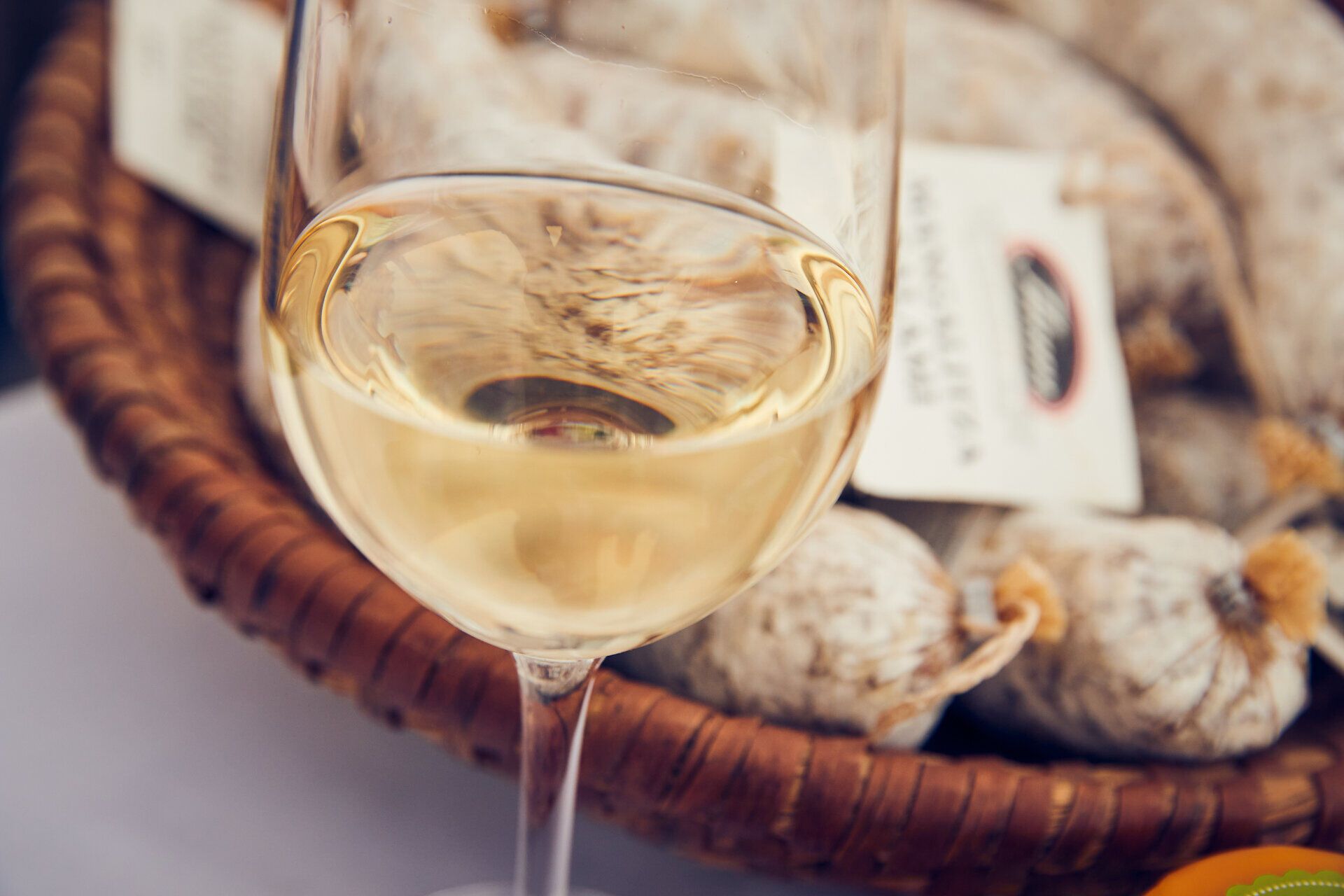 Ein elegantes Glas Weißwein schimmert im Licht und verspricht Genuss und Entspannung. Im Hintergrund liegen köstliche Wurstwaren, die die Vorfreude auf eine kulinarische Entdeckungstour wecken. Hier, inmitten der Weinregion, wird jeder Schluck zum Erlebnis.