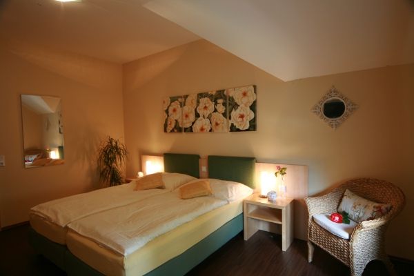 Gemütliches Schlafzimmer mit Doppelbett, Rattan-Sessel, Nachttisch und Blumenbild an der Wand.