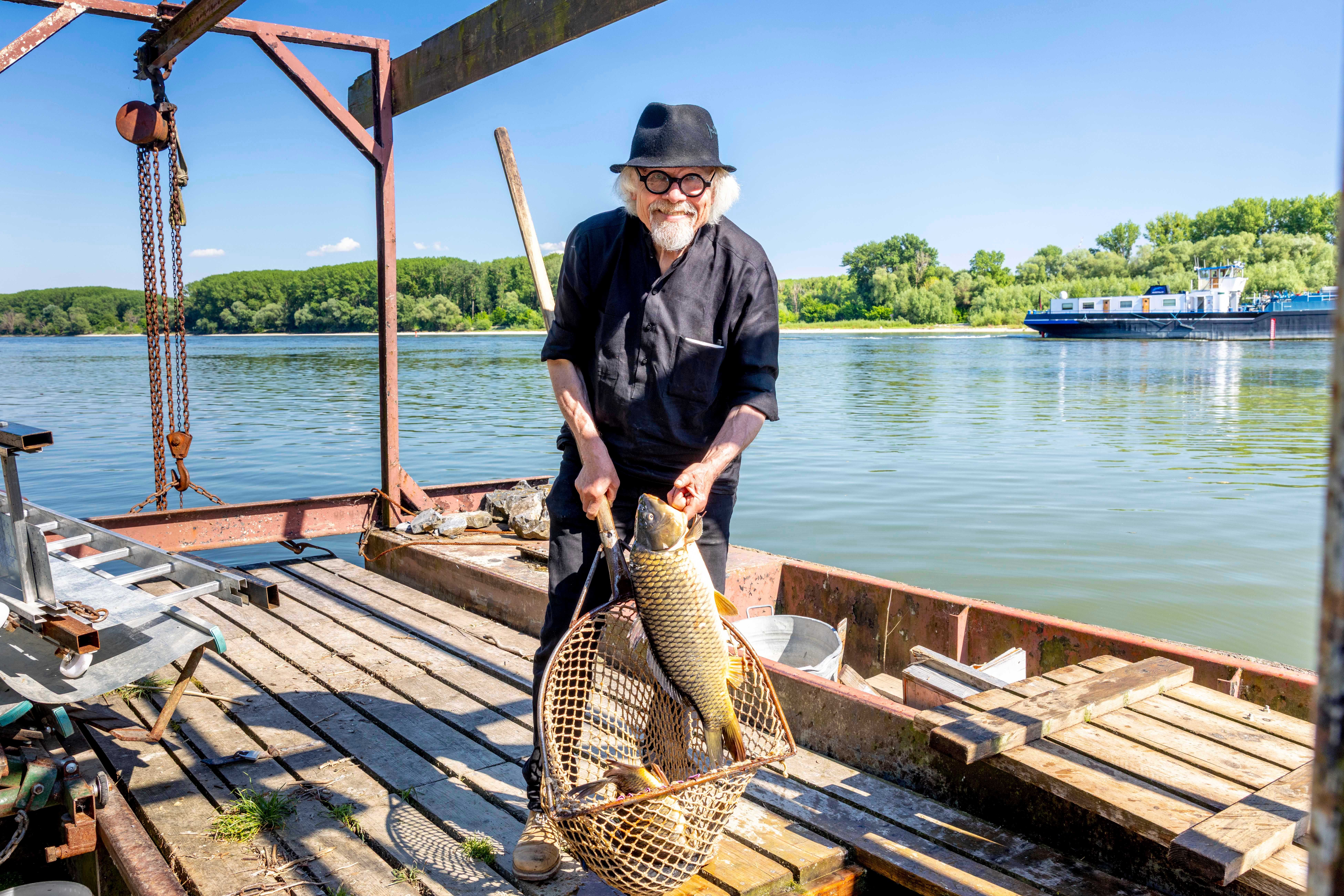 Ein Mann mit Hut hält einen großen Fisch in einem Netz auf einem Holzsteg am Fluss (Donau).