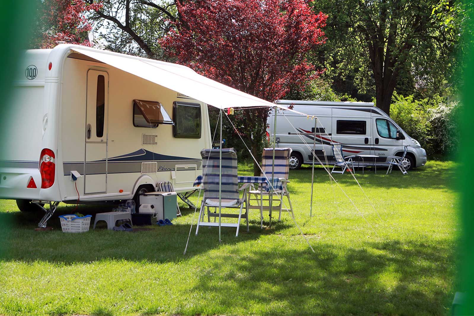Wohnwagen und Wohnmobil auf einem grünen Campingplatz mit Stühlen und Tisch unter einer Markise.