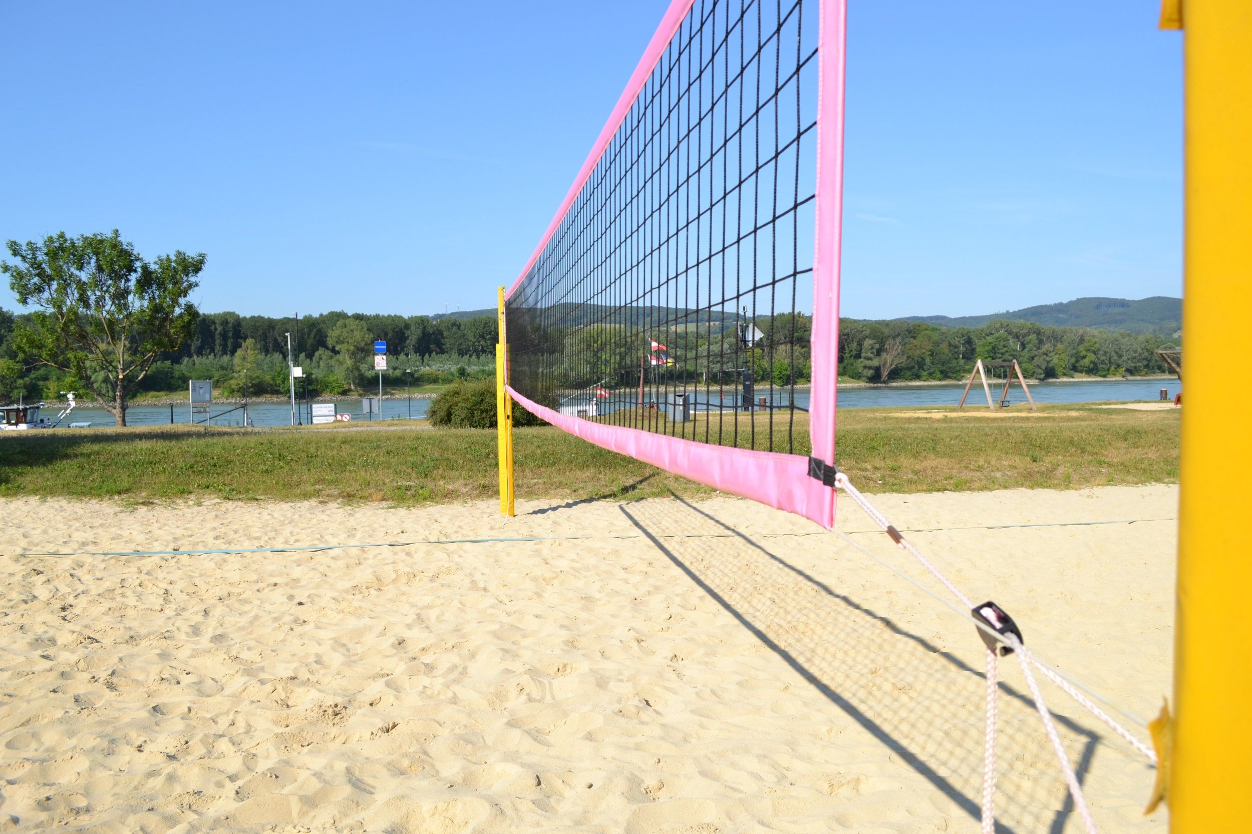 Beachvolleyballplatz mit Netz, Sand und Fluss im Hintergrund.