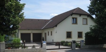 Ein modernes Einfamilienhaus mit braunem Dach und hellem Anstrich, umgeben von Bäumen und einem Zaun.