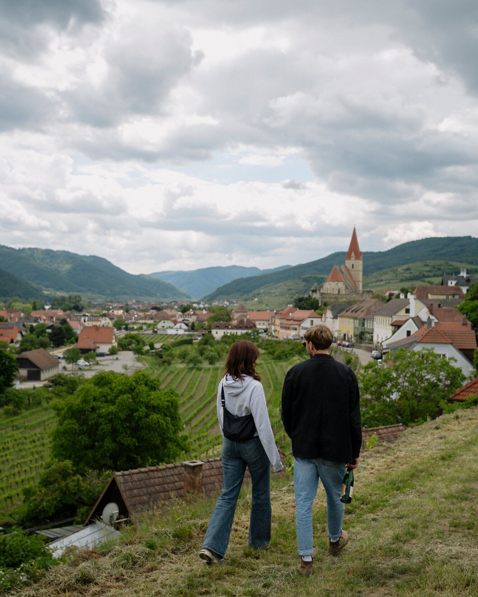 Zwei Personen gehen auf einem Hangweg oberhalb eines Ortes; vor ihnen liegen Dächer und Weingärten, im Hintergrund ragt der Kirchturm von Weißenkirchen vor wolkigem Himmel auf.