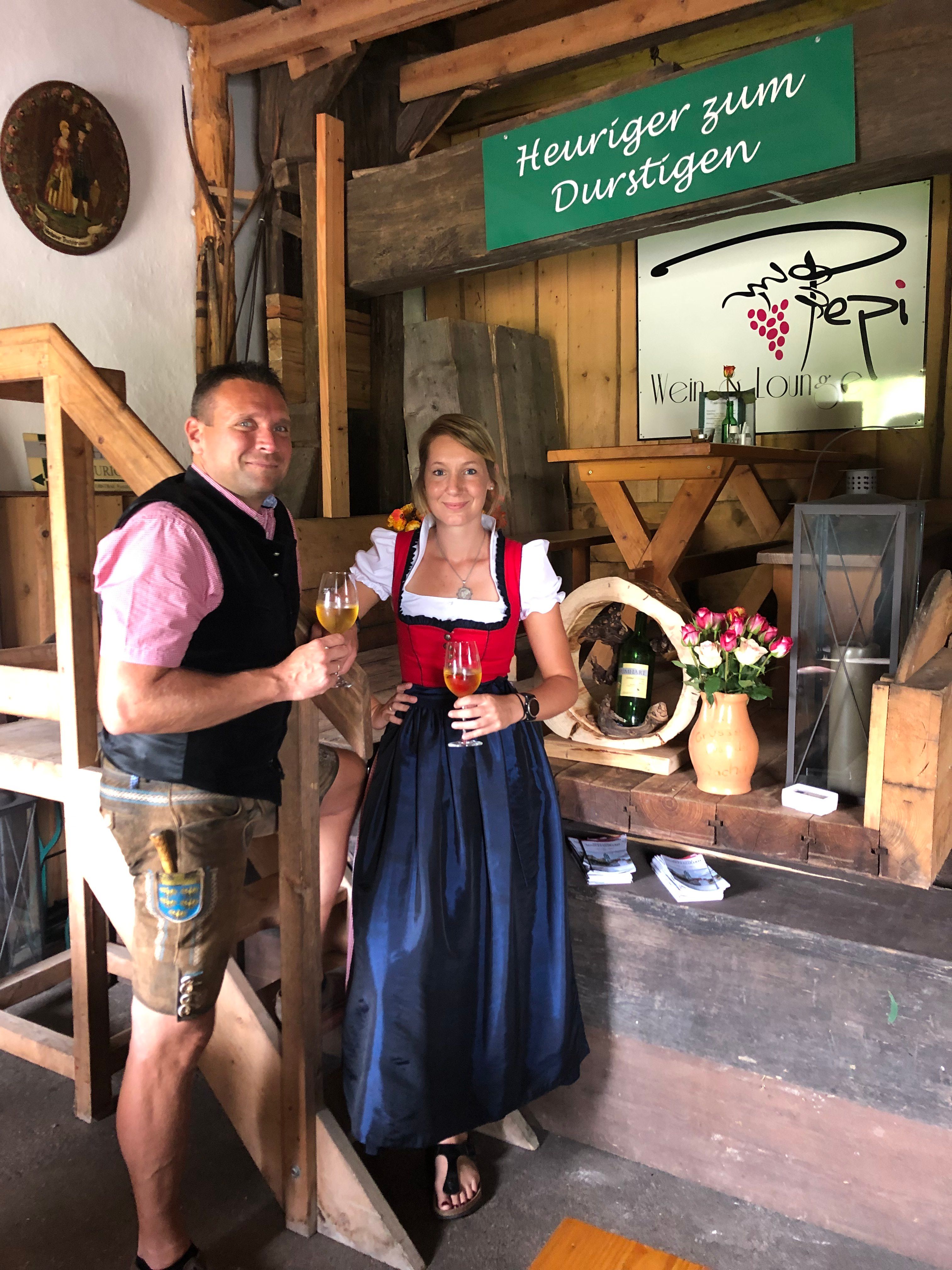 Zwei Personen in traditioneller Kleidung bei einem Heurigen, mit Weingläsern in der Hand.