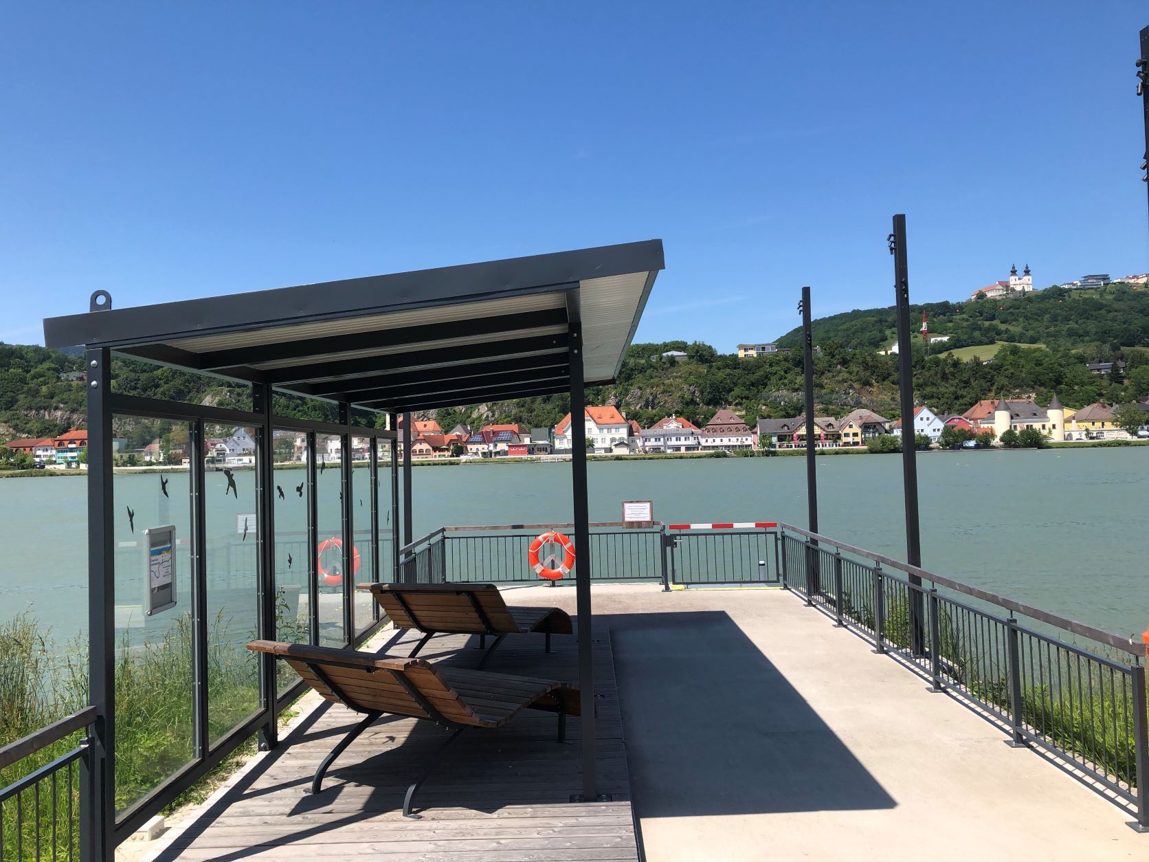 Anlegestelle mit Überdachung und Liegen, Fluss im Hintergrund, Basilika auf Hügel.