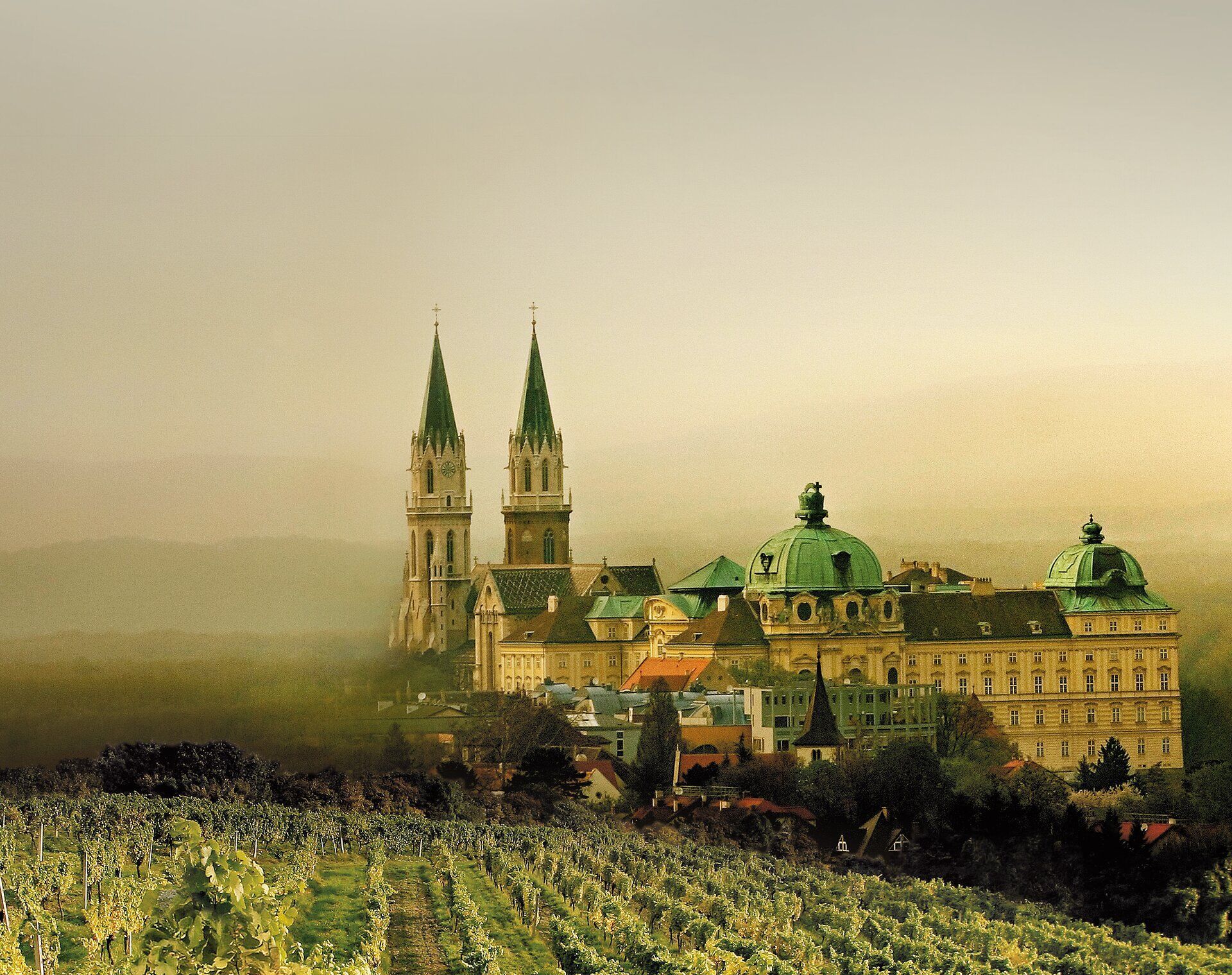 Die sanften Hügel umgeben das majestätische Stift, dessen markante Türme in den Himmel ragen. Ein Hauch von Geschichte und Spiritualität erfüllt die Luft, während die Weinreben im Vordergrund die Schönheit der Umgebung unterstreichen.