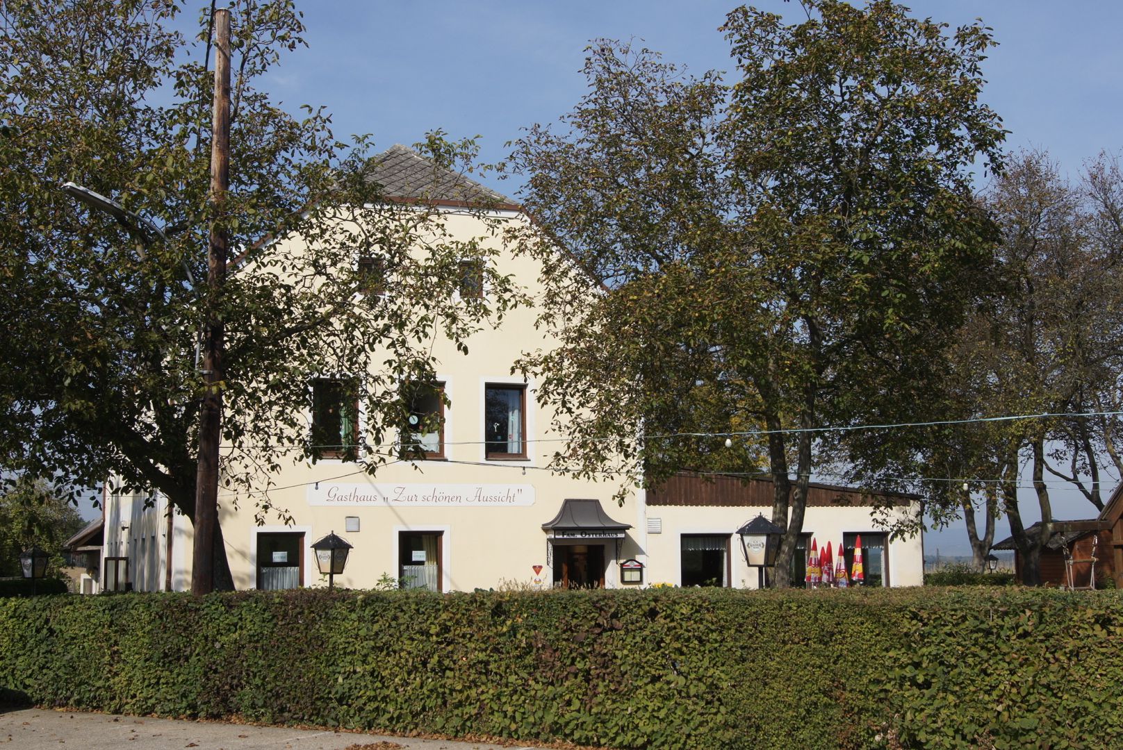 Ein gelbes Gasthaus mit der Aufschrift 'Gasthaus Zur schönen Aussicht', umgeben von Bäumen und Hecken.