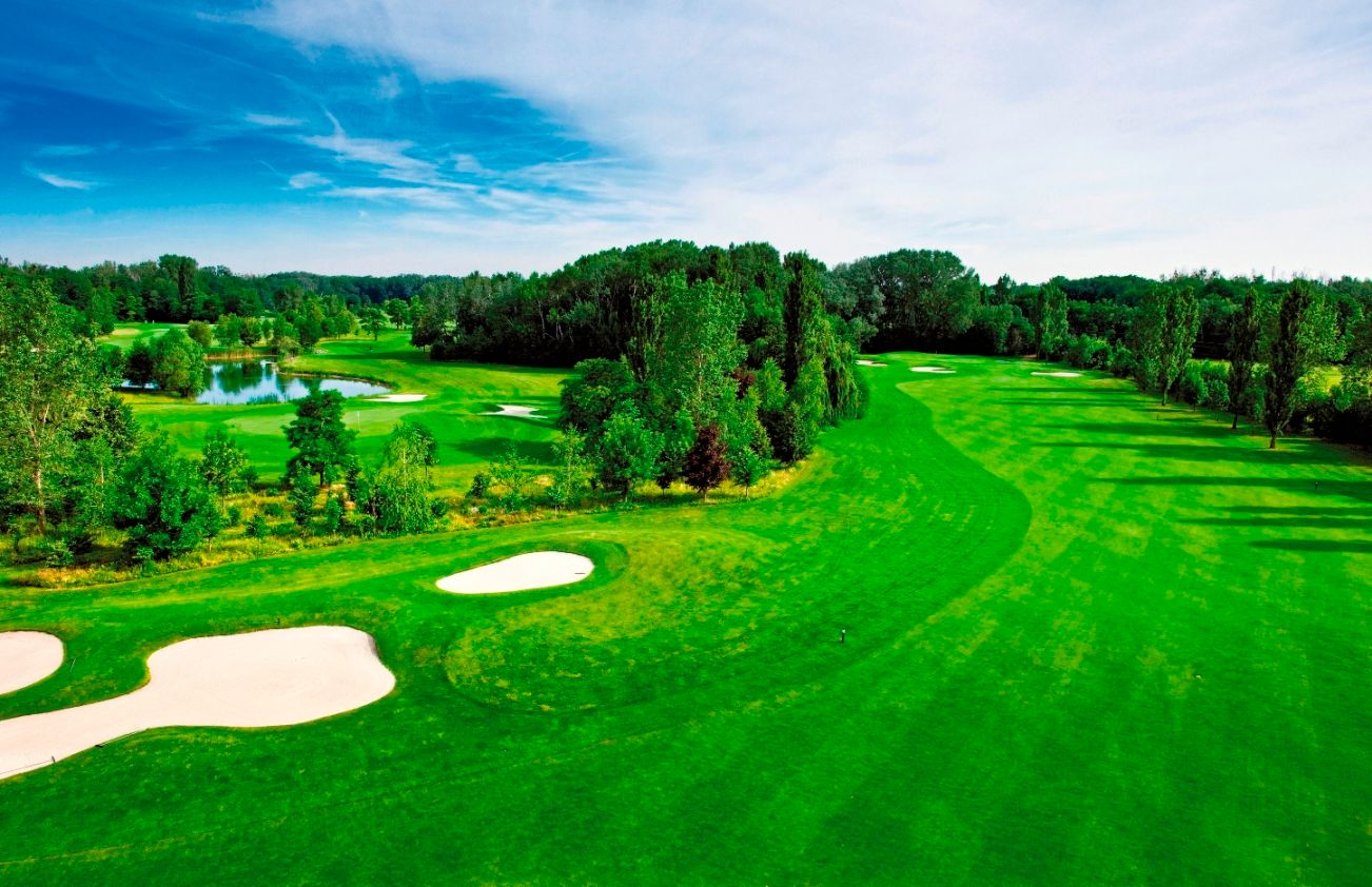 Zöld golfpálya bunkerekkel és tóval, fákkal körülvéve, kék ég alatt.