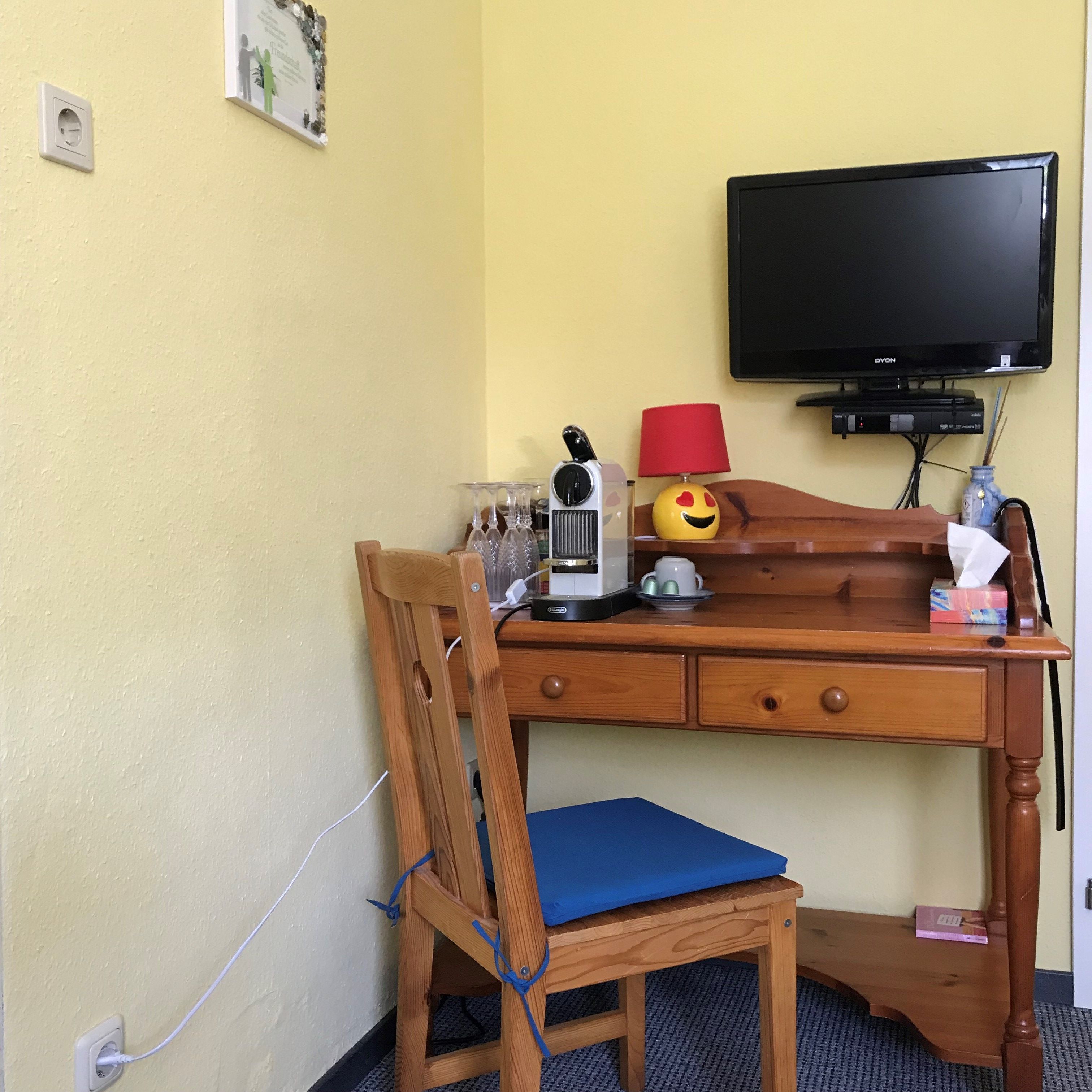 Ein kleiner Holztisch mit einer Kaffeemaschine, Gläsern, einer Lampe und einem Fernseher an der Wand.