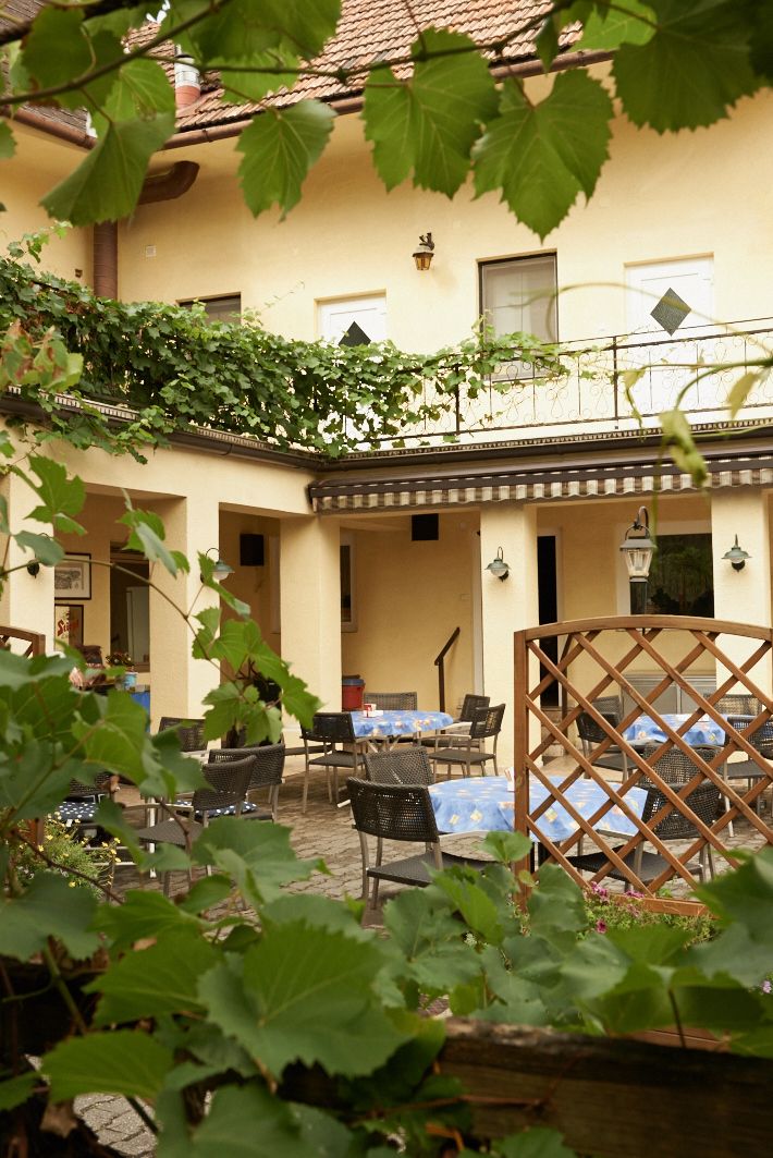 Ein gemütlicher Gastgarten mit Tischen und Stühlen, umgeben von grünen Blättern und einem gelben Gebäude im Hintergrund.