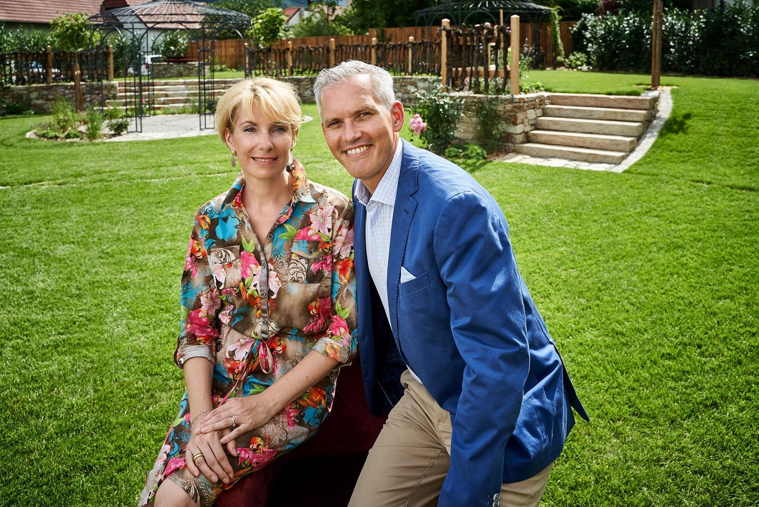 Familie Wildeis sitzet auf einer Wiese, lächelnd in die Kamera. Der Mann trägt ein blaues Sakko, die Frau ein buntes Kleid.