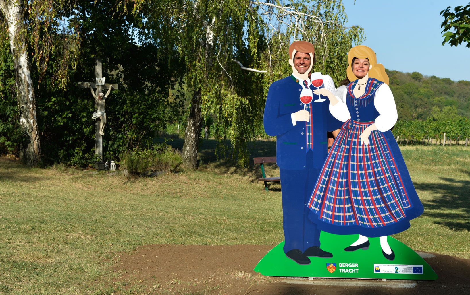 Zwei Personen posieren hinter einer Pappfigur in traditioneller Tracht mit Weingläsern. Im Hintergrund ein Kruzifix und Bäume.