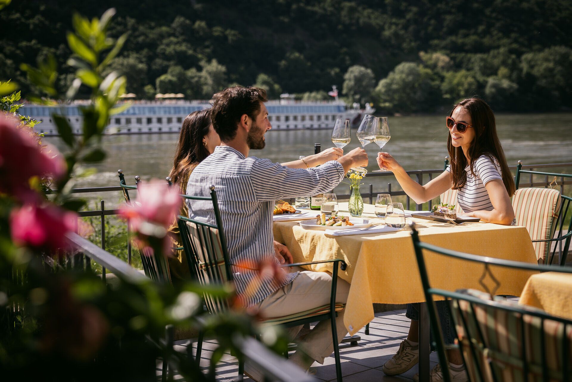 Zwei Damen und ein Herr sitzen an einem gedeckten Tisch und prosten sich zu.