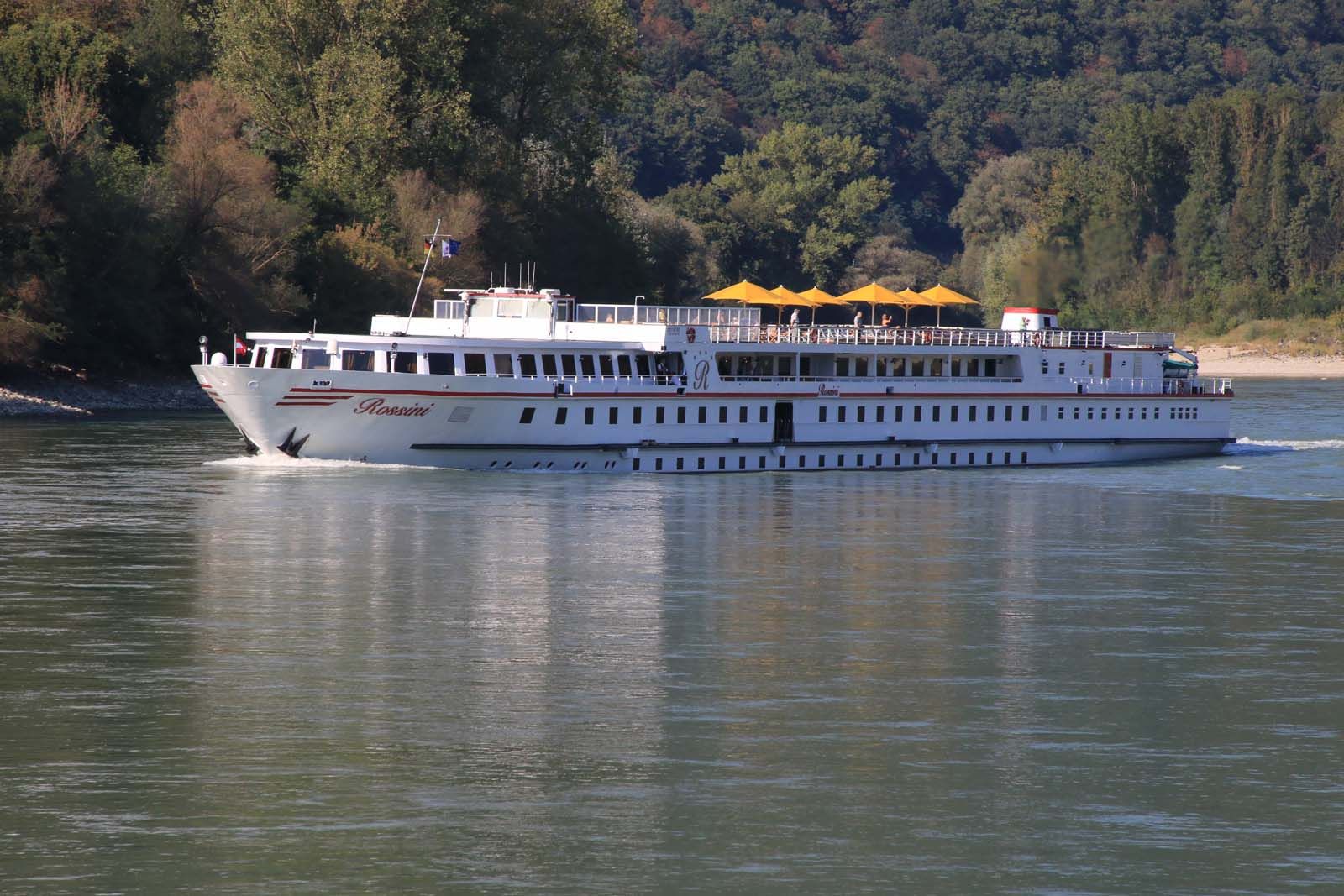 Ein Flusskreuzfahrtschiff namens Rossini fährt auf der Donau, umgeben von bewaldeten Ufern.