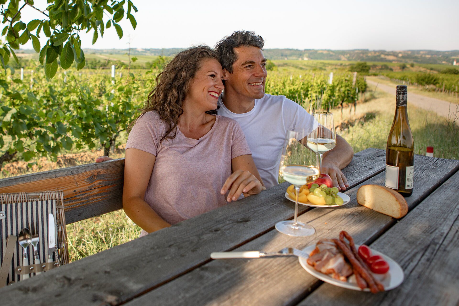 Paar sitzt entspannt an einem Holztisch im Weingarten mit Weinflaschen.