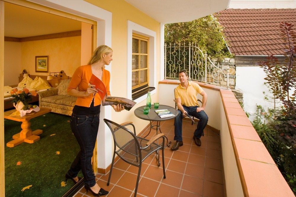 Ein Mann sitzt auf einem Balkon, eine Frau kommt aus dem Zimmer mit einem Magazin. Tisch mit Getränken, gemütliche Atmosphäre.