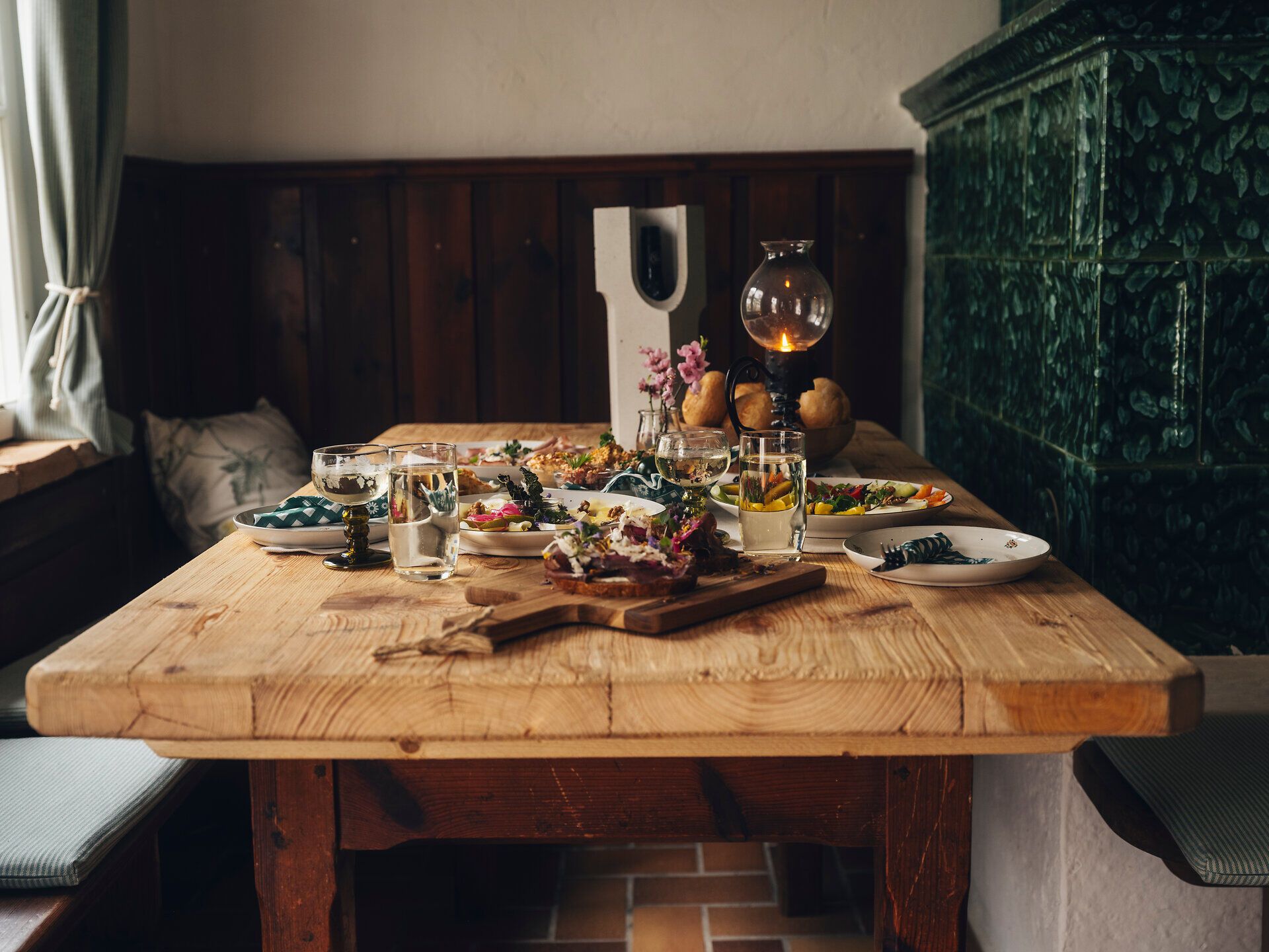 Rustikaler Holztisch mit angerichteten Speisen, Brot, Gläsern und Blumen in einer gemütlichen Stube; im Hintergrund eine Holzvertäfelung und ein grüner Kachelofen.