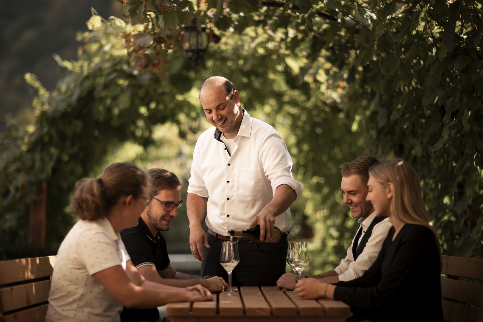 Ein Mann schenkt Wein für vier Gäste an einem Tisch im Freien ein, umgeben von grünen Weinreben.
