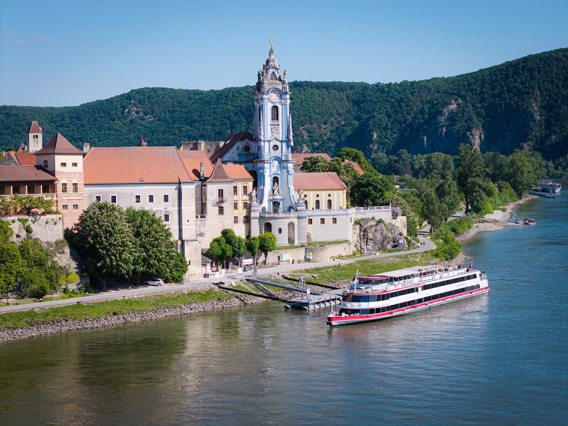 Die sanften Hügel umarmen das malerische Ufer der Donau, während die historische Architektur des Ortes in der warmen Sonne erstrahlt. Ein gemütliches Schiff gleitet über das Wasser und lädt dazu ein, die Schönheit dieser einzigartigen Landschaft zu entdecken.