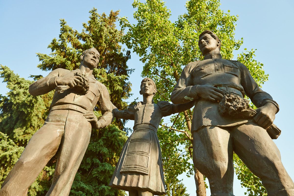 Drei Bronzestatuen, zwei Männer und eine Frau, stehen nebeneinander vor Bäumen.
