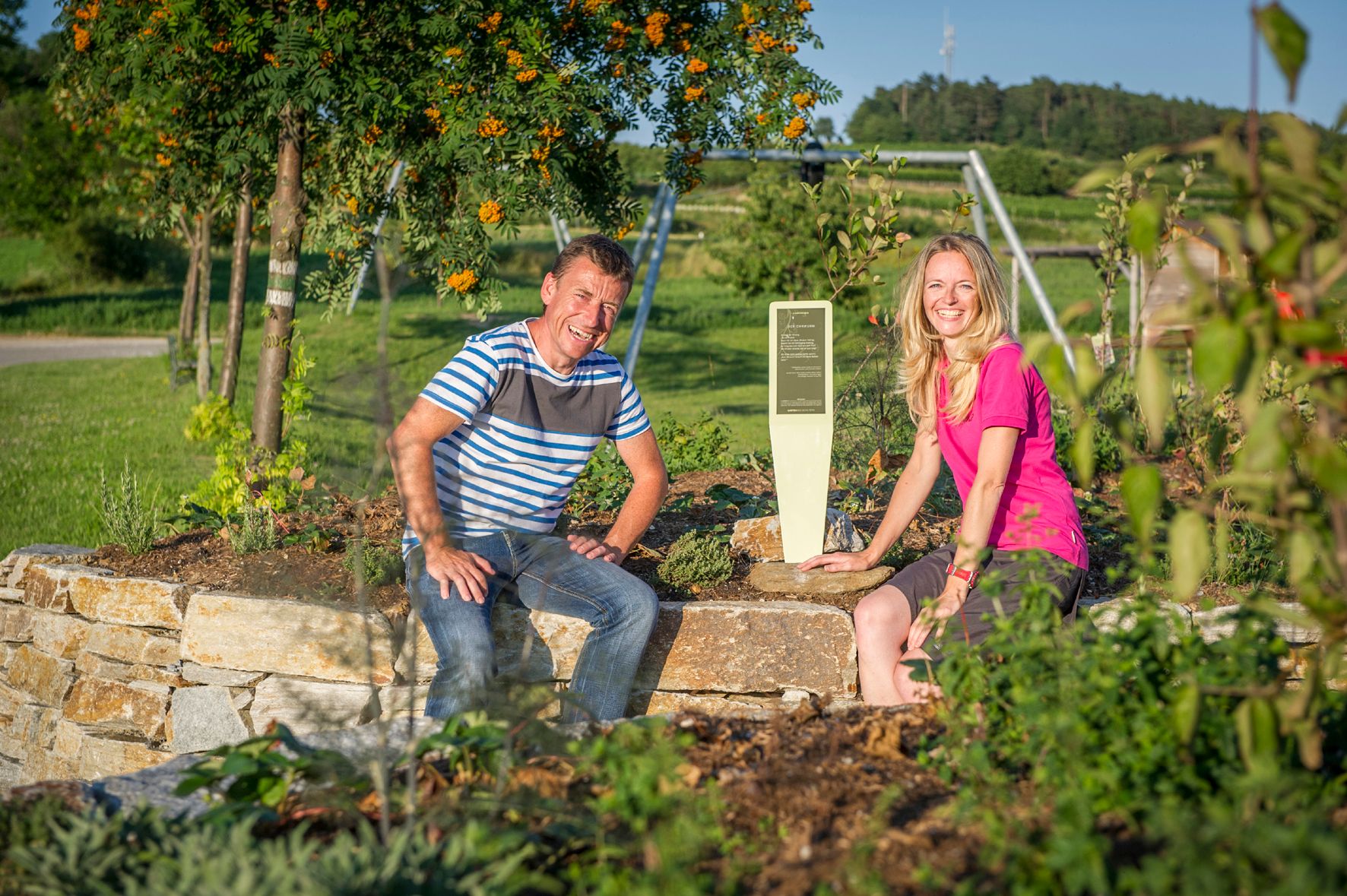 Zwei Personen sitzen auf einer Steinmauer in einem Garten mit Pflanzen und einem Schild.