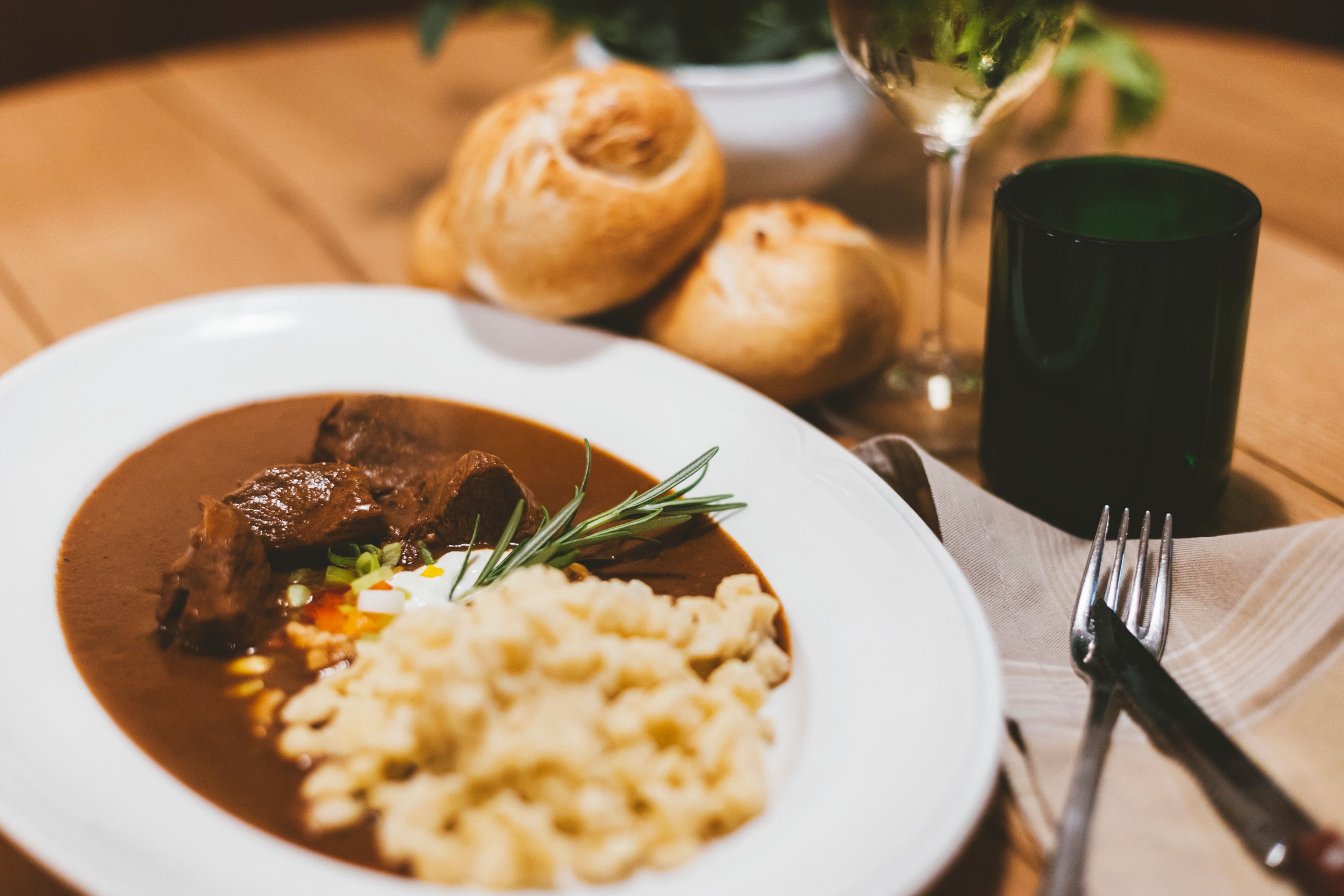 Teller mit Gulasch und Spätzle, Brot und Weinglas im Hintergrund.