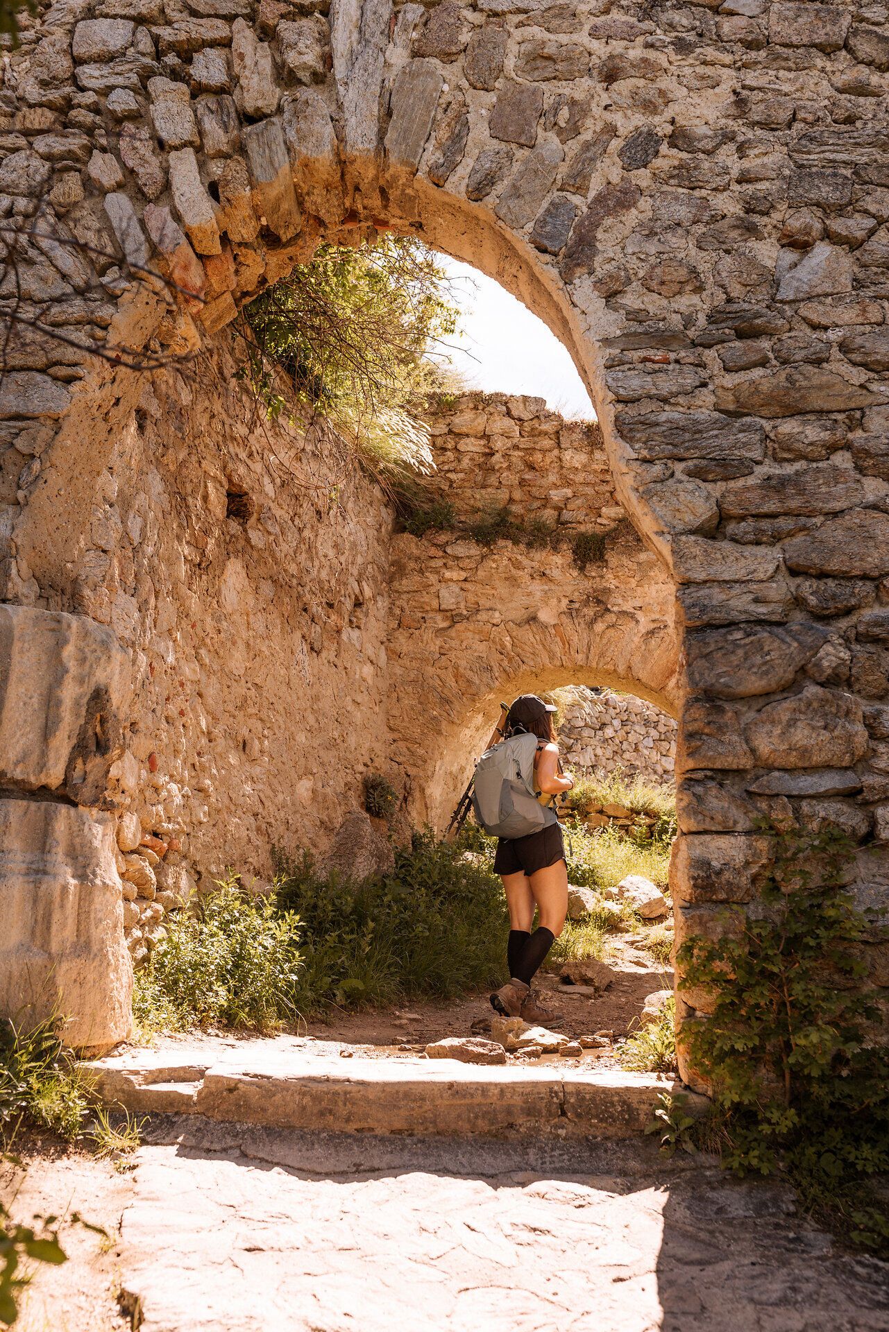 Eine Frau mit Wanderrucksack geht unter einer gemauerten Ruine hindurch.
