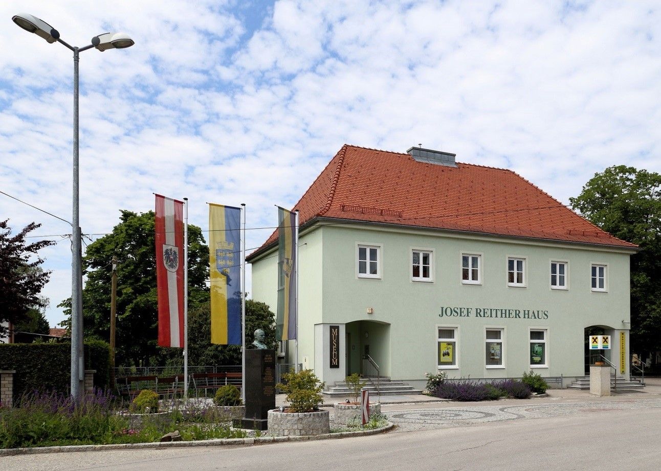 Josef Reither Haus mit Flaggen und Büste im Vordergrund.
