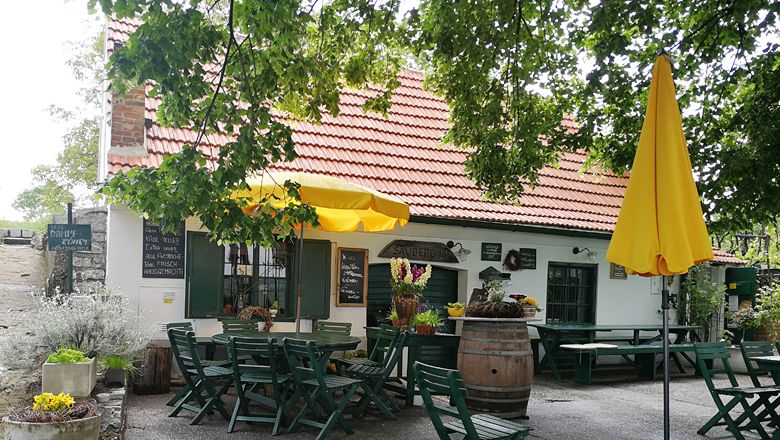 Gemütlicher Gastgarten mit gelbem Sonnenschirm und Holztischen vor einem Gebäude mit rotem Ziegeldach.
