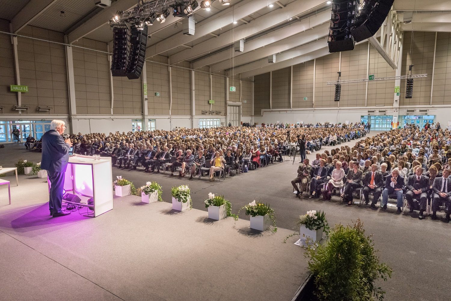 A speaker addresses a large audience in an exhibition hall.