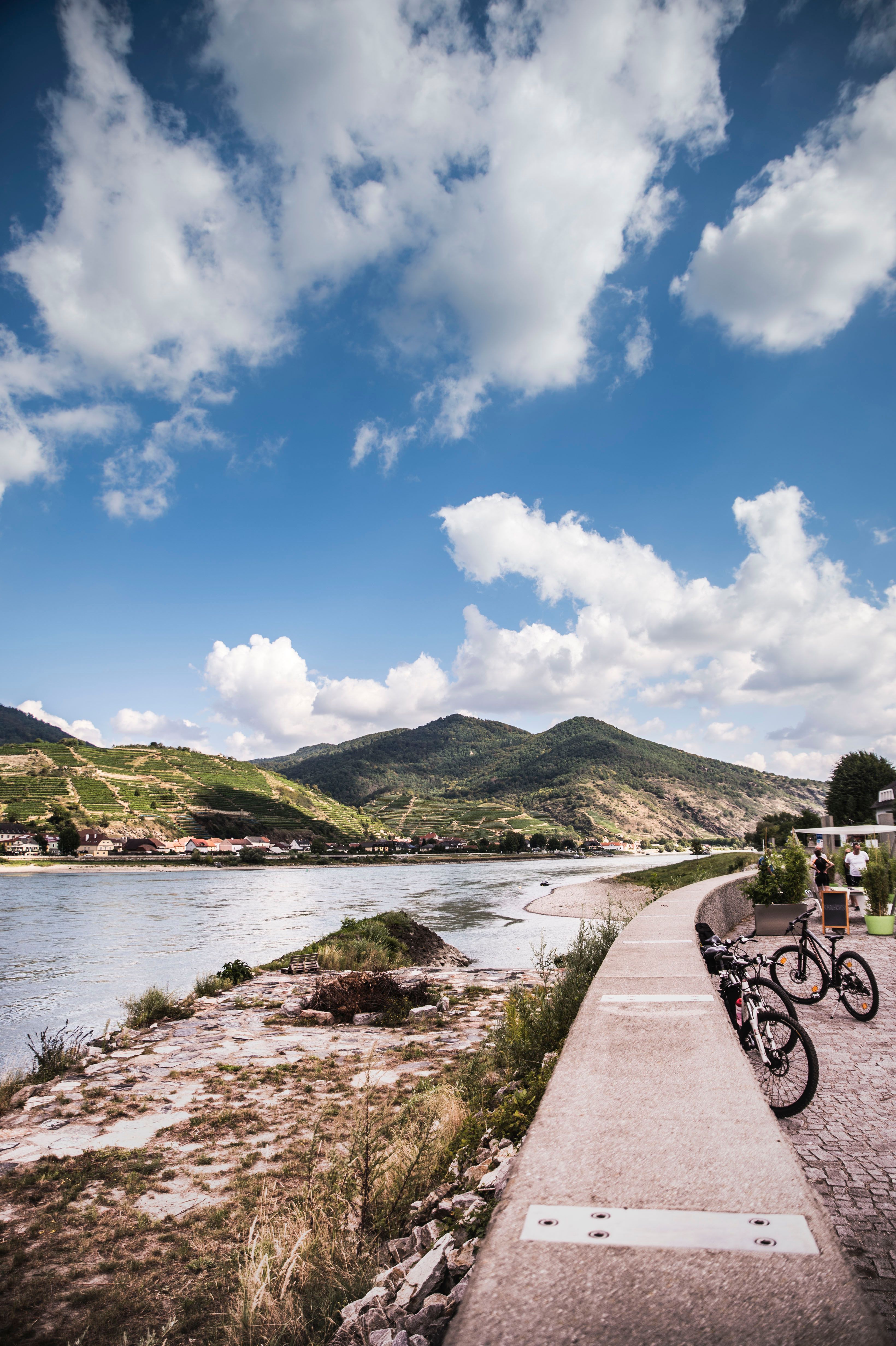 River landscape with flood protection wall, bicycles and hills in the background.