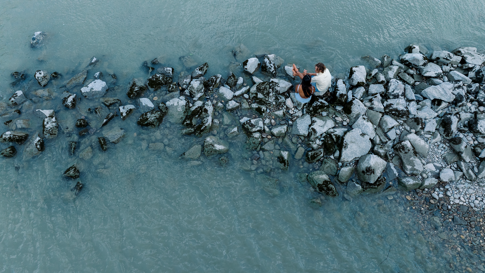 Luftaufnahme: Zwei Personen sitzen auf einer schmalen Stein- und Schotterbank, umgeben von graublauem Flusswasser.
