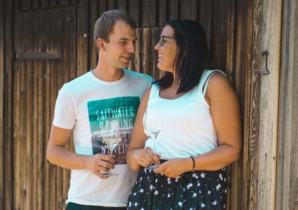 Ein Mann und eine Frau stehen lächelnd vor einer Holzwand und halten Weingläser.