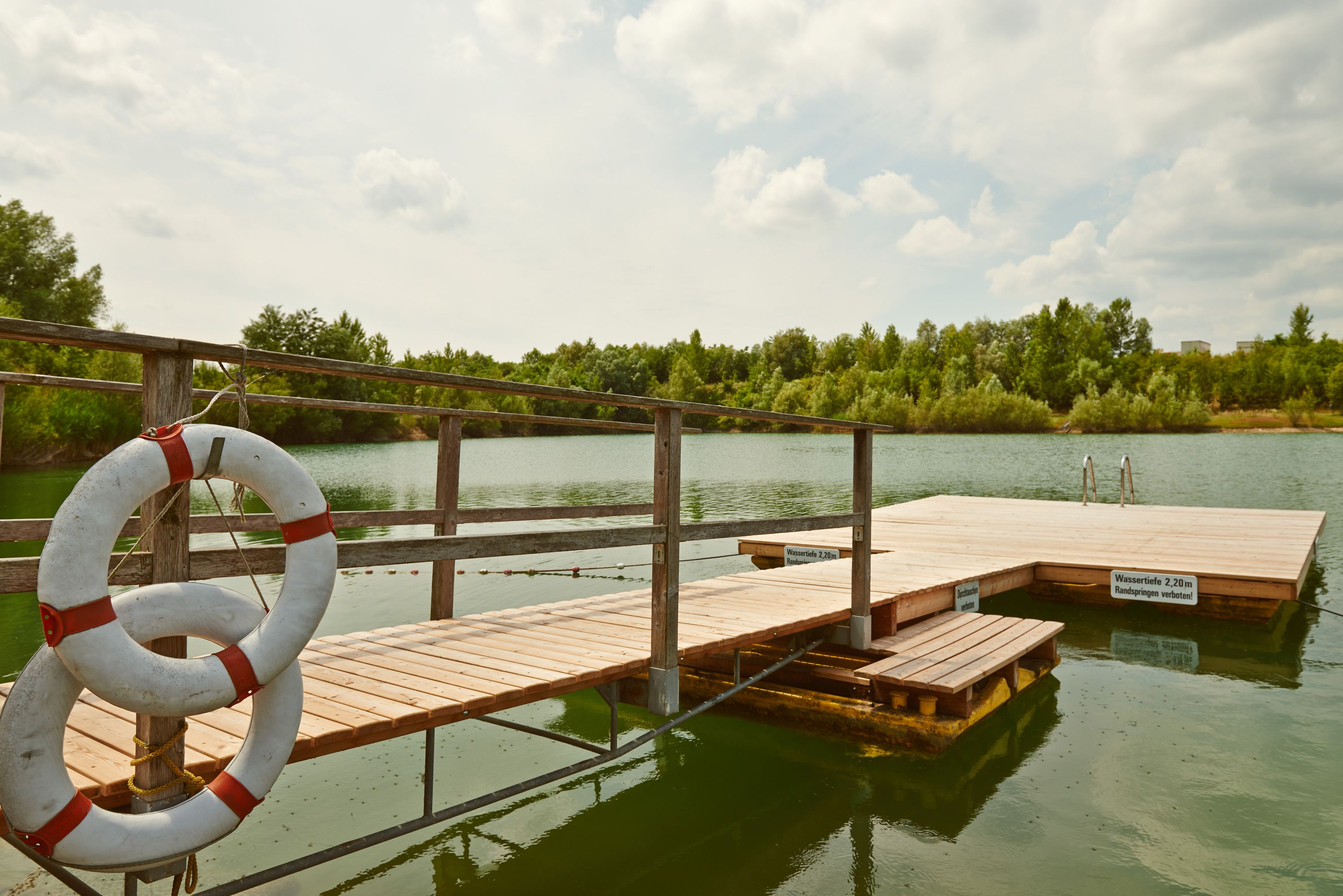 Ein hölzerner Steg mit Rettungsringen an einem ruhigen See, umgeben von Bäumen.