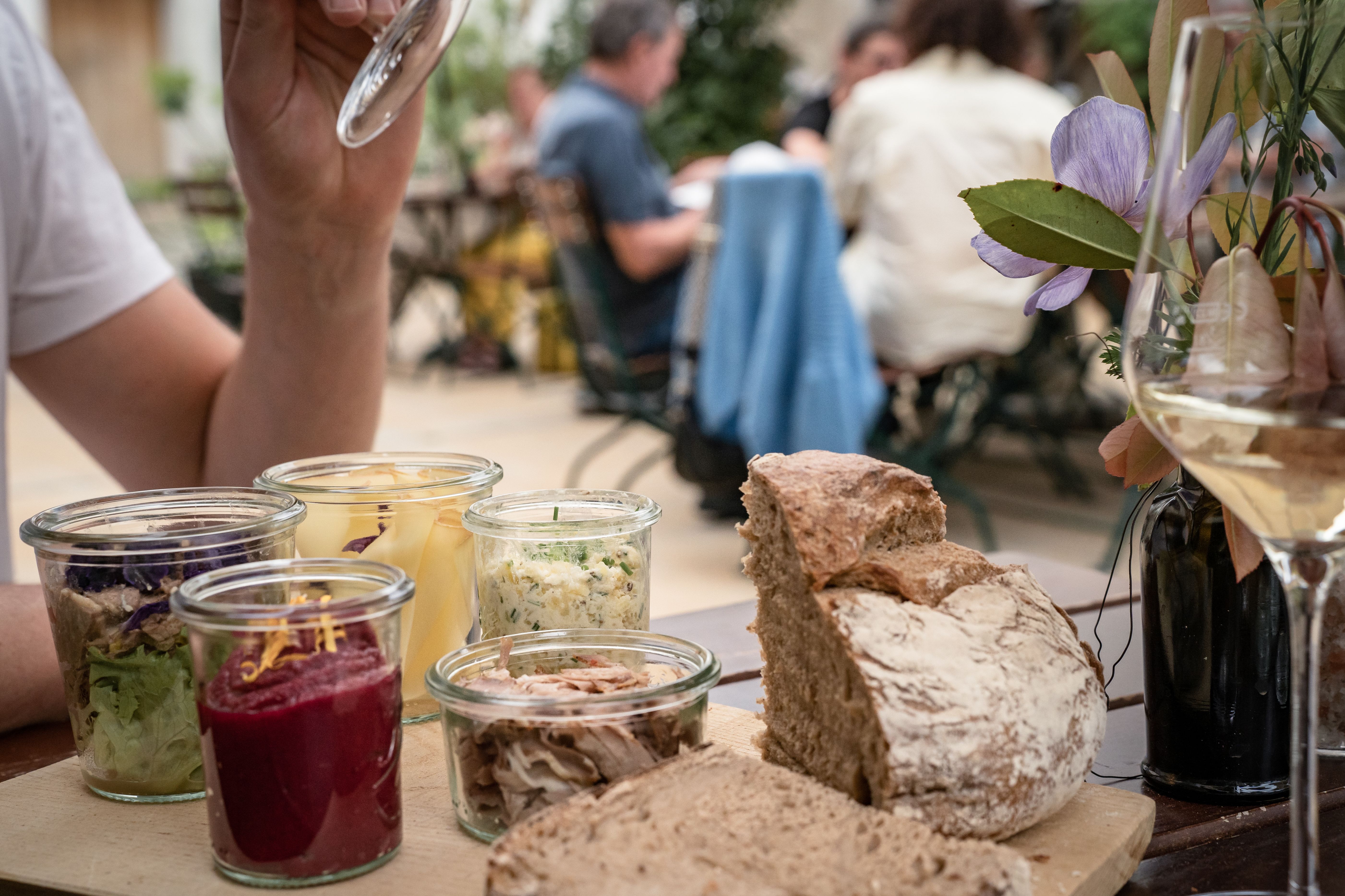 Holzbrett mit aufgeschnittenem Bauernbrot und mehreren kleinen Gläsern mit Aufstrichen und Salaten auf einem Gartentisch. Daneben ein Glas Weißwein und eine Blumenvase, im Hintergrund sitzen Gäste unscharf in einem Gastgarten.