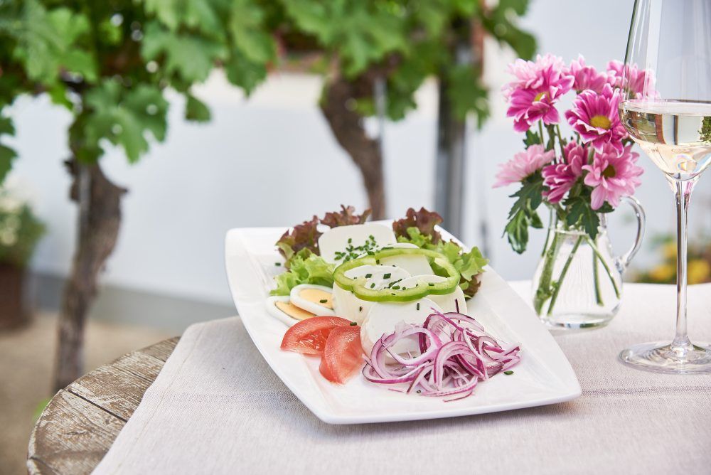 Ein Teller mit Käse, Zwiebeln, Tomaten und Salat auf einem Tisch mit Blumen und einem Glas Weißwein.