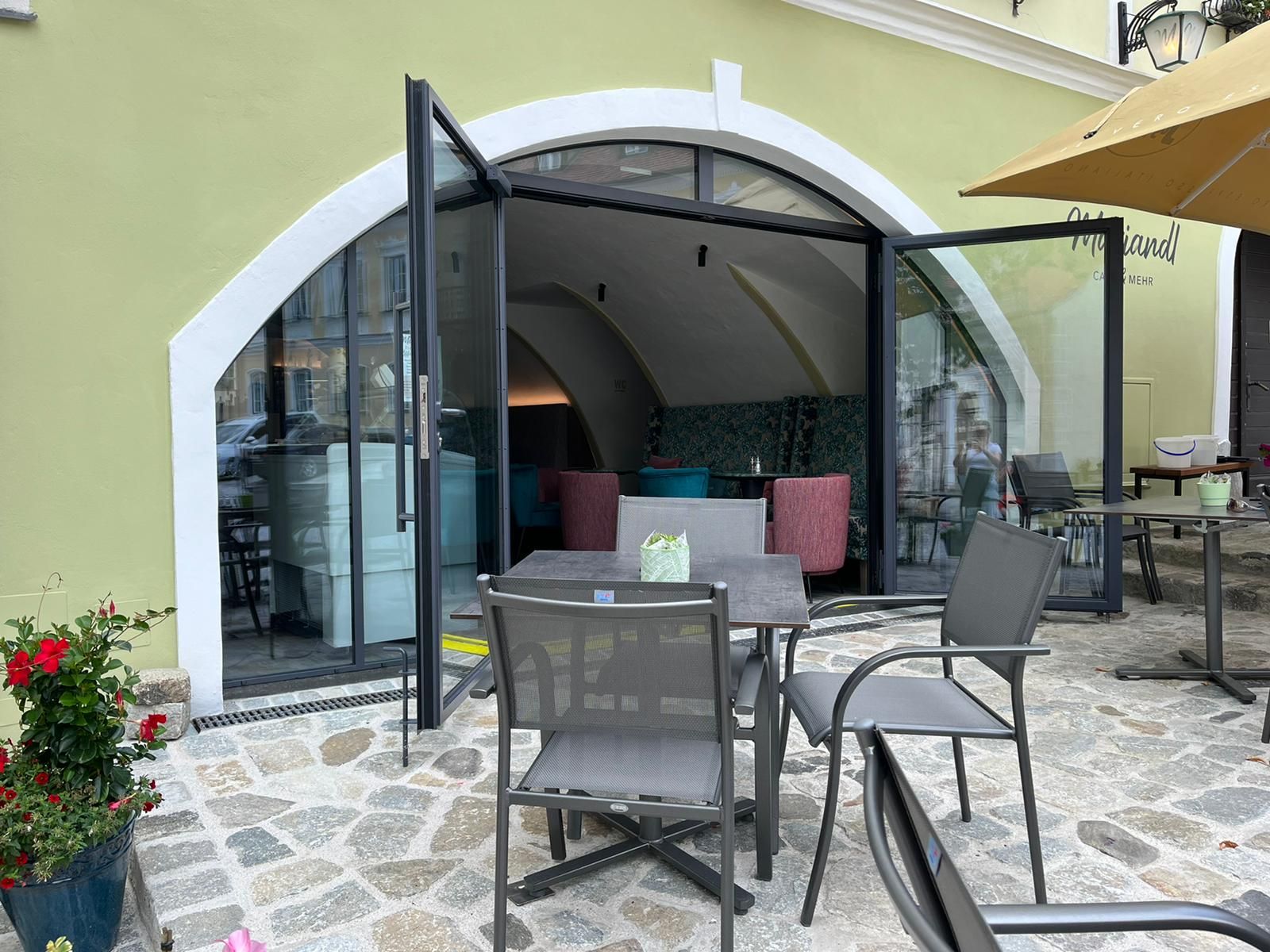 Außenansicht eines Cafés mit offenen Glastüren und Sitzgelegenheiten im Freien.