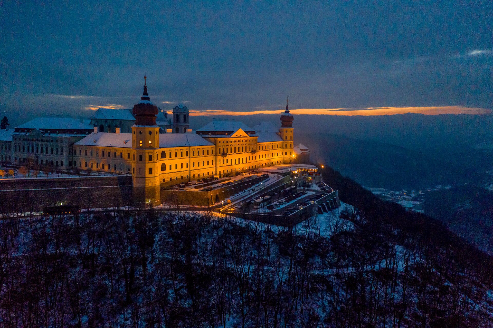 In der winterlichen Dämmerung erstrahlt das Kloster Göttweig in warmem Licht und schafft eine magische Atmosphäre. Die schneebedeckten Dächer und die sanften Hügel im Hintergrund laden dazu ein, die besinnliche Adventszeit in vollen Zügen zu genießen.