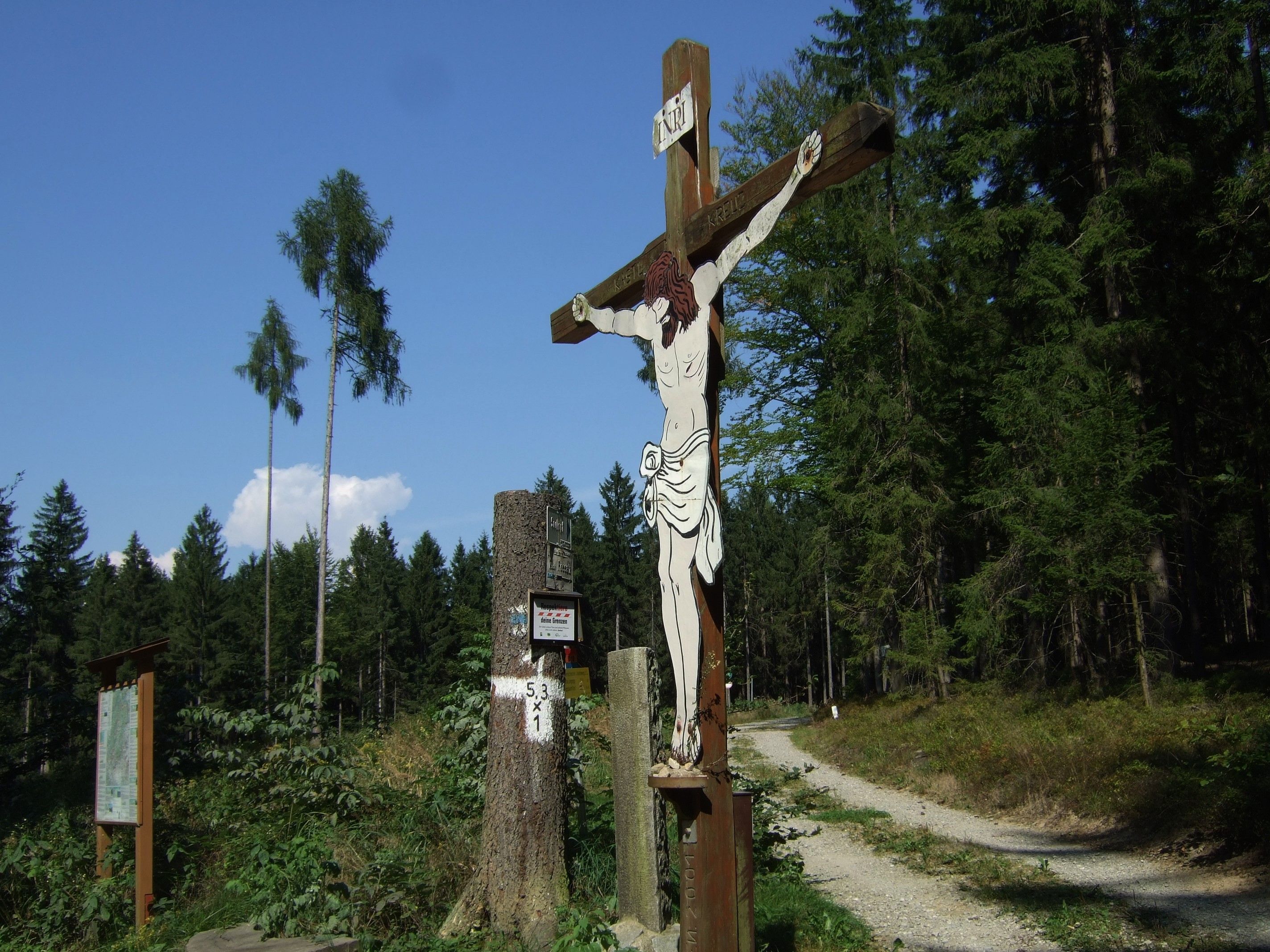 Dřevěný kříž na okraji lesa s cestou v pozadí.