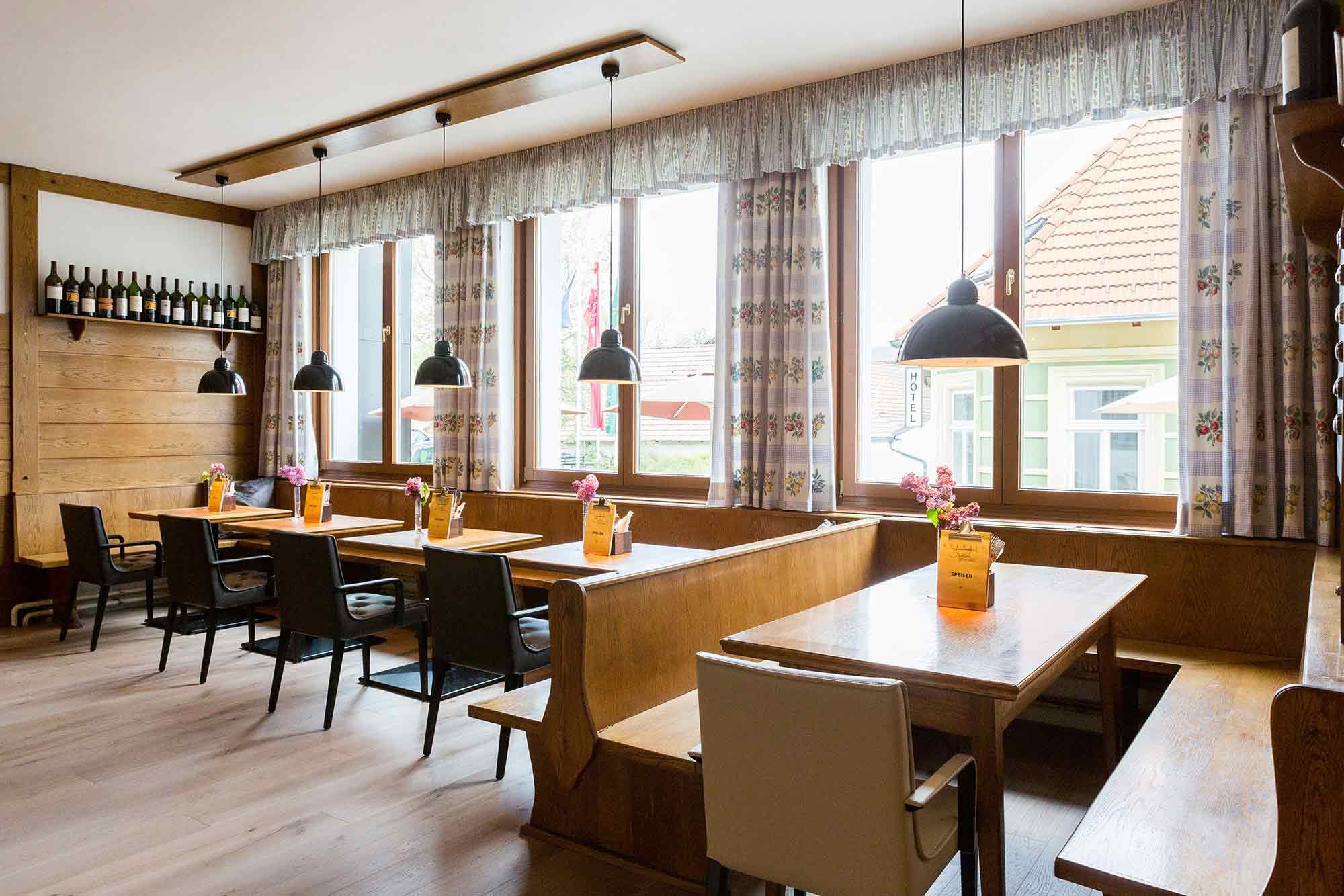 Gemütliches Gastzimmer mit Holztischen, Stühlen und Bänken, dekoriert mit Blumen und Speisekarten. Große Fenster mit Vorhängen und hängenden Lampen.