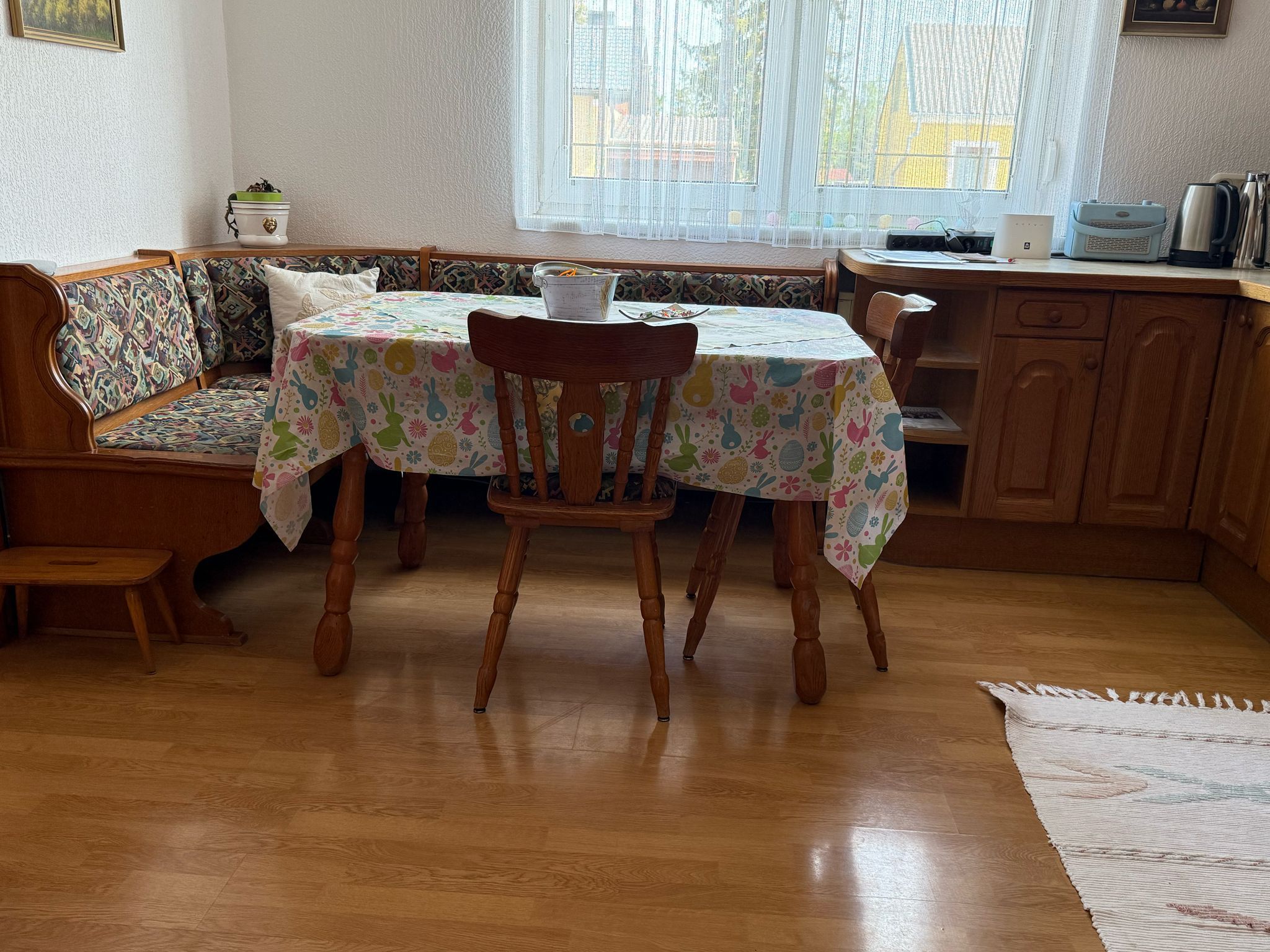 Essbereich mit Holztisch, Stühlen und Eckbank, bunte Tischdecke, Fenster im Hintergrund.