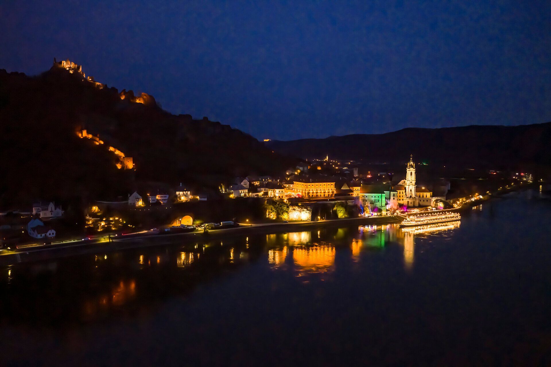 Nächtlicher Blick auf das festlich beleuchtete Dürnstein an der Donau, mit reflektierenden Lichtern, ruhiger Flusslandschaft und historischer Kirche im Advent.