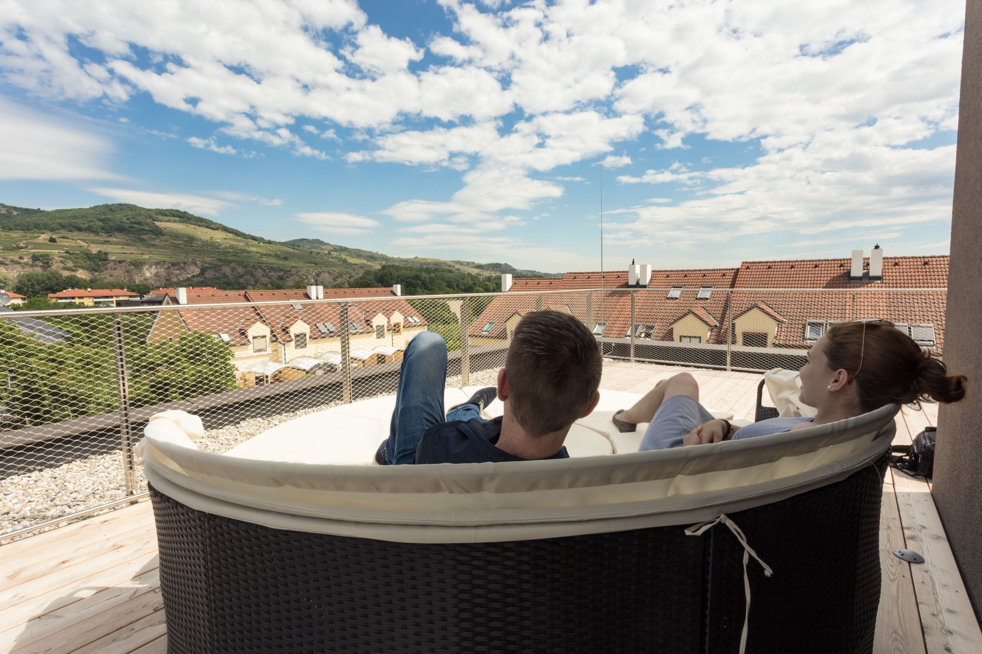 Zwei Personen entspannen auf einer Terrasse mit Blick auf Dächer und Hügel.