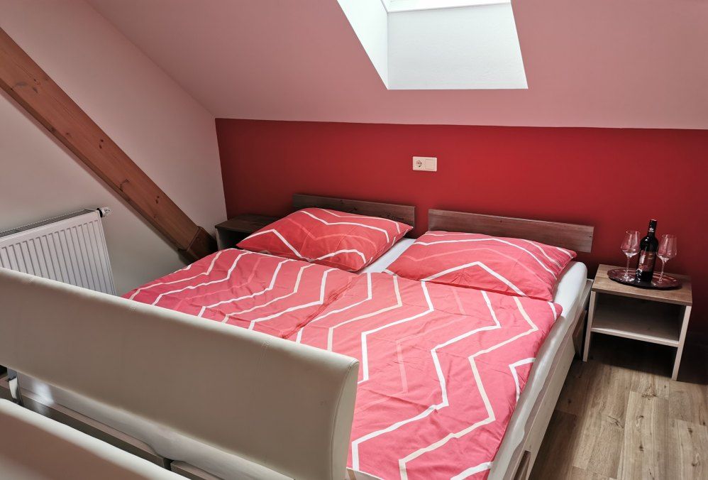 Schlafzimmer mit roter Wand, Doppelbett mit roter Bettwäsche, Dachfenster und Nachttisch mit Weinflasche und Gläsern.