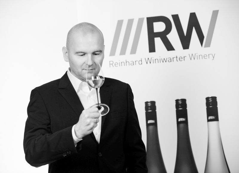Öltönyös férfi egy pohár bort szagolgat egy "Reinhard Winiwarter Pincészet" feliratú transzparens előtt.