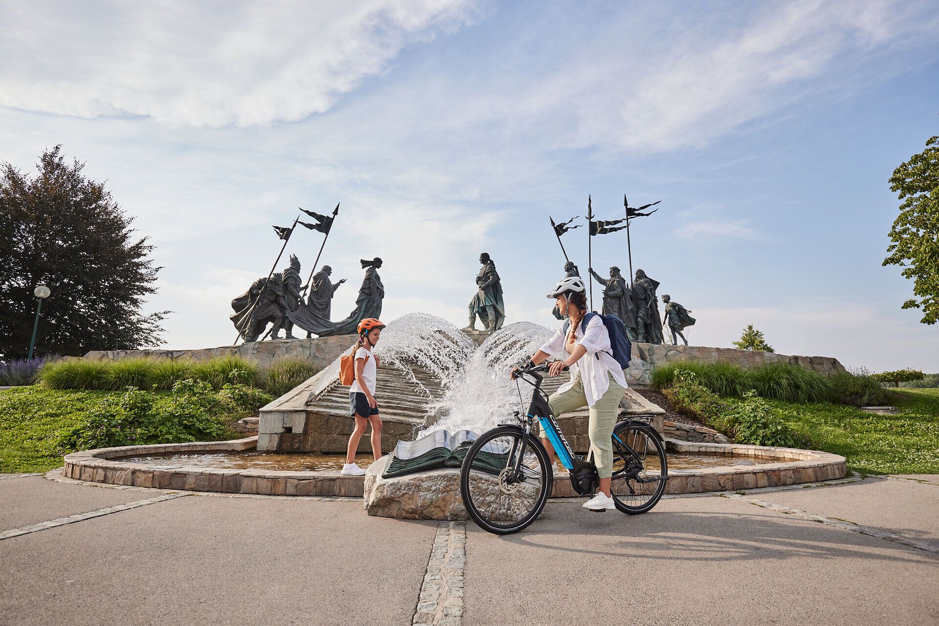 Eine Familie genießt einen sonnigen Tag in der Nähe eines sprudelnden Brunnens, umgeben von beeindruckenden Statuen. Die frische Luft und die lebendige Atmosphäre laden dazu ein, die Umgebung mit dem Fahrrad zu erkunden und die Schönheit der Natur zu erleben.