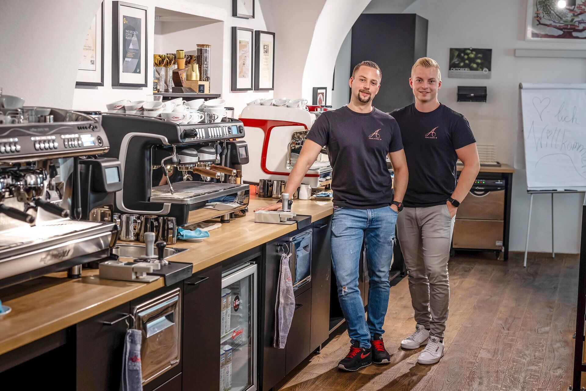 In einem einladenden Café, umgeben von modernem Design und der warmen Atmosphäre frisch gebrühten Kaffees, stehen zwei Baristas bereit, um ihre Leidenschaft für das Handwerk zu teilen. Die Düfte von frisch gemahlenen Bohnen und köstlichen Backwaren laden dazu ein, einen Moment der Entspannung zu genießen und die Kunst des Kaffeegenusses zu erleben.