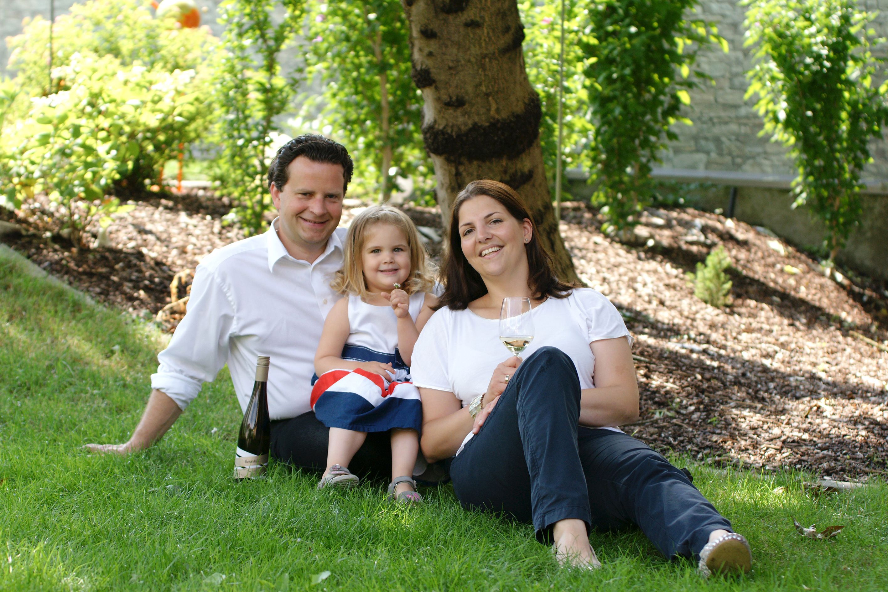 Familie sitzt auf einer Wiese unter einem Baum mit Weinflasche und Glas.