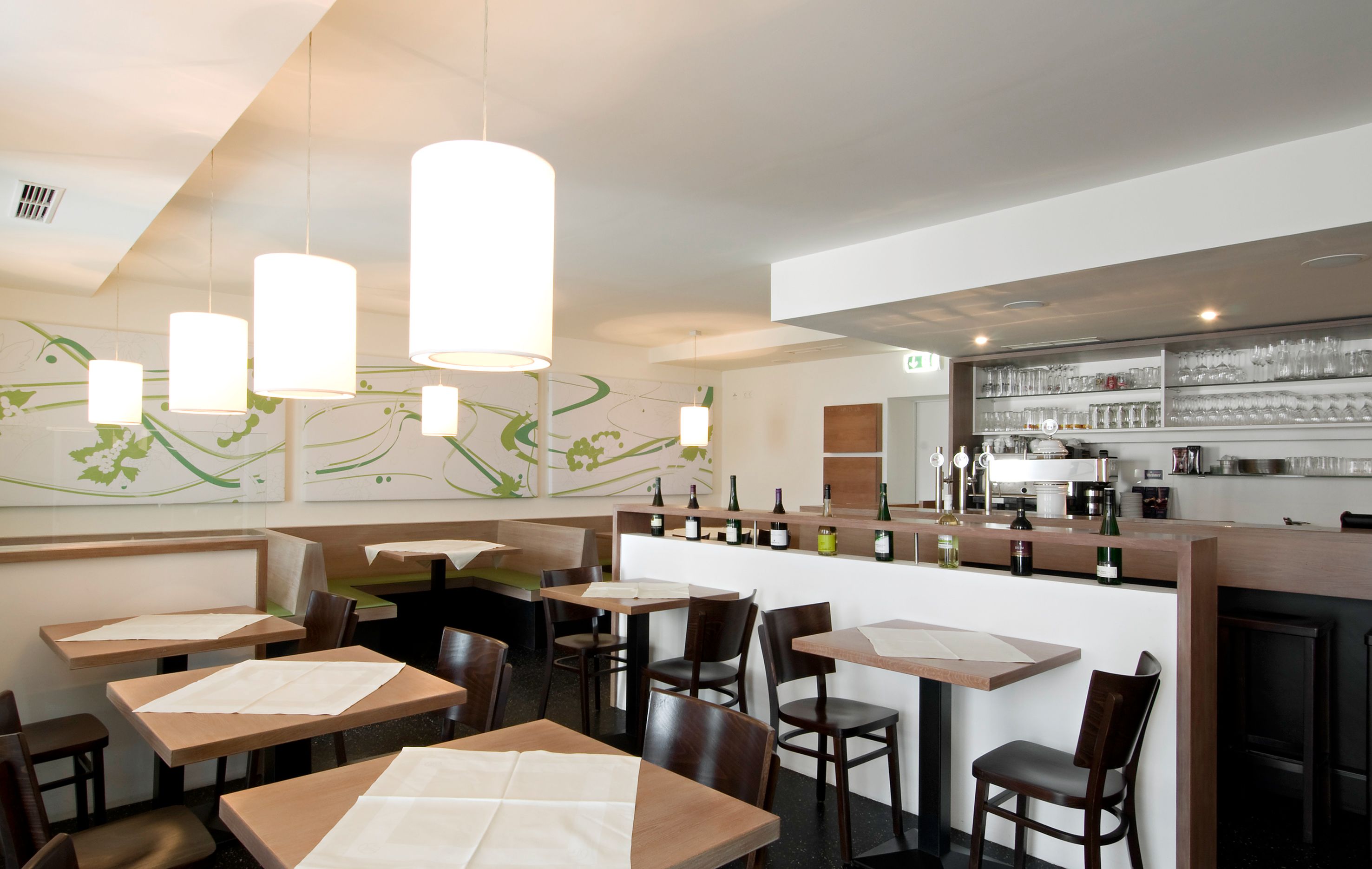 Innenansicht eines modernen Restaurants mit Holztischen, Stühlen und einer Bar mit Flaschen und Gläsern.