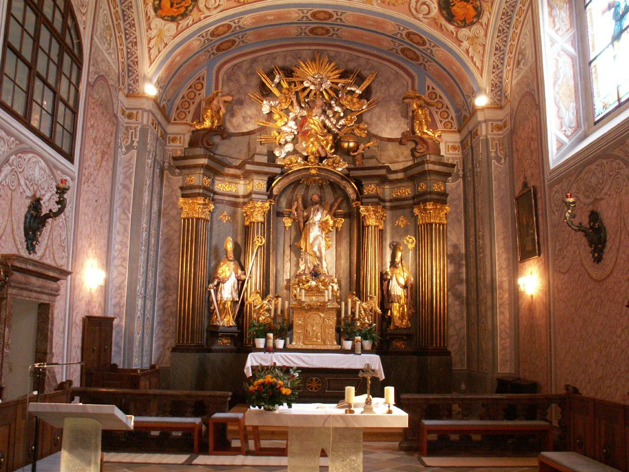 Innenansicht einer barocken Kirche mit reich verziertem Hochaltar, goldenen Säulen und Heiligenfiguren, davor ein schlichter Altar mit Kerzen.